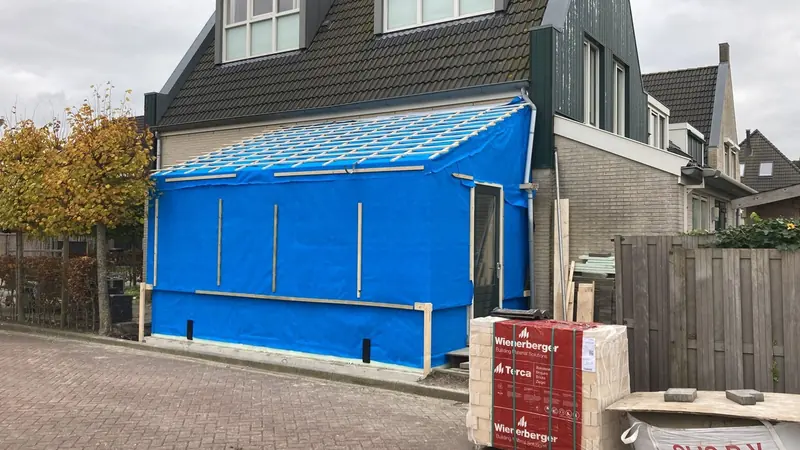 Aanbouw in Sijbekarspel tijdens de bouw onder blauw zeil, met pallet bakstenen ervoor