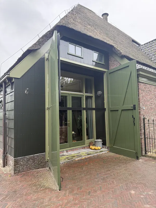 Open groene deeldeuren voor een glazen pui onder de rieten kap, stolpboerderij in Opmeer