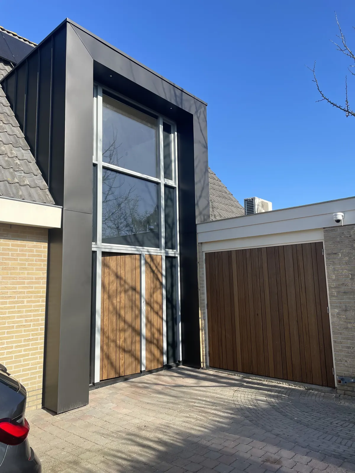 Hoge glazen pui in donkergrijze omlijsting met houten deuren, naast de houten garagedeur van een woning in Hoorn