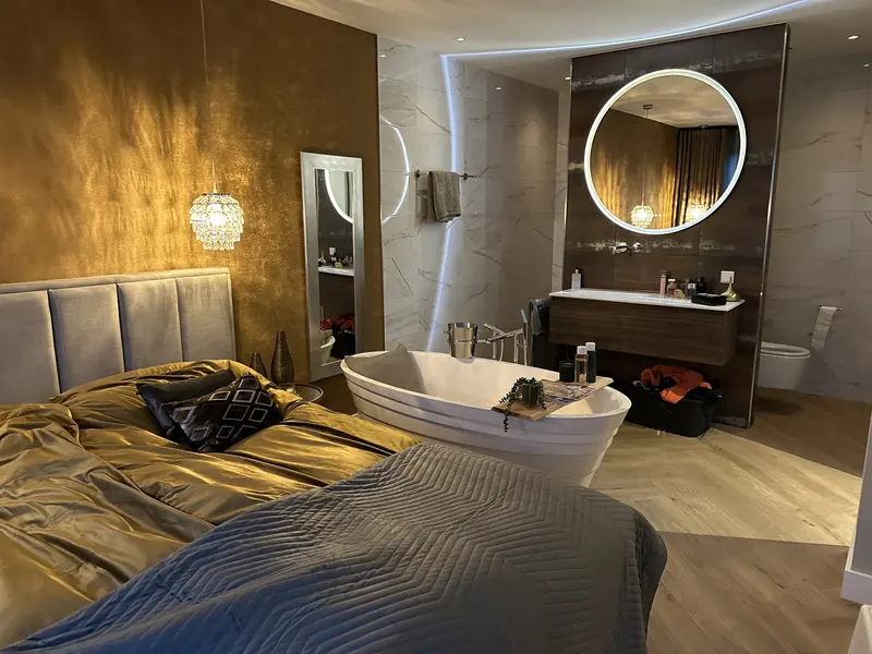 Open badkamer met vrijstaand bad, wastafelmeubel en ronde verlichte spiegel naast de slaapkamer, Oosthuizen
