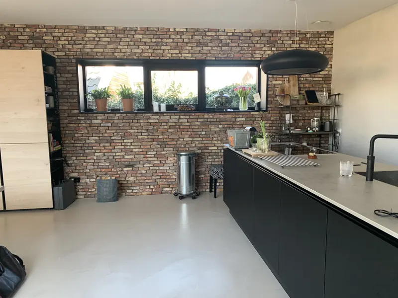Keuken met zwart eiland, gietvloer en een wand van baksteen rond een breed raam, Broek op Langedijk